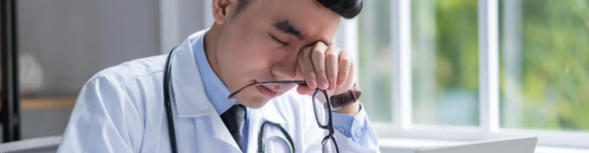 Healthcare provider sitting at a desk looking fatigued while reviewing work on a laptop, representing burnout and career stress