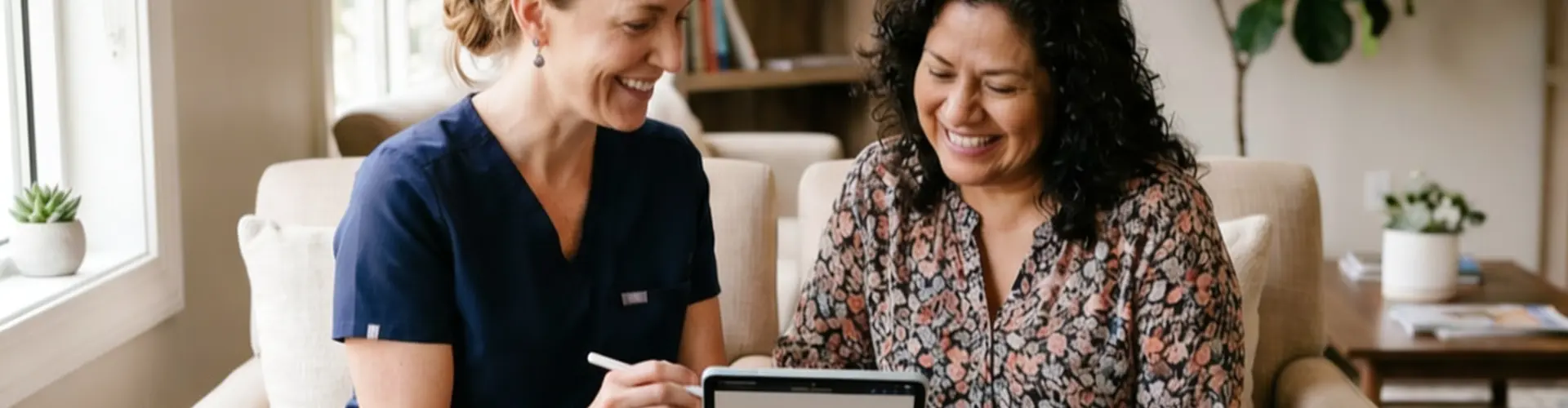 Nurse practitioner in scrubs sitting beside a patient in a comfortable, home-like setting, reviewing health information together on a tablet during a collaborative coaching session.